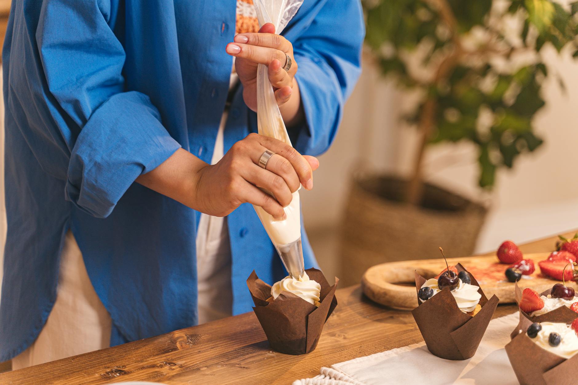 Person using piping bag to decorate cupcakes in a kitchen setting.