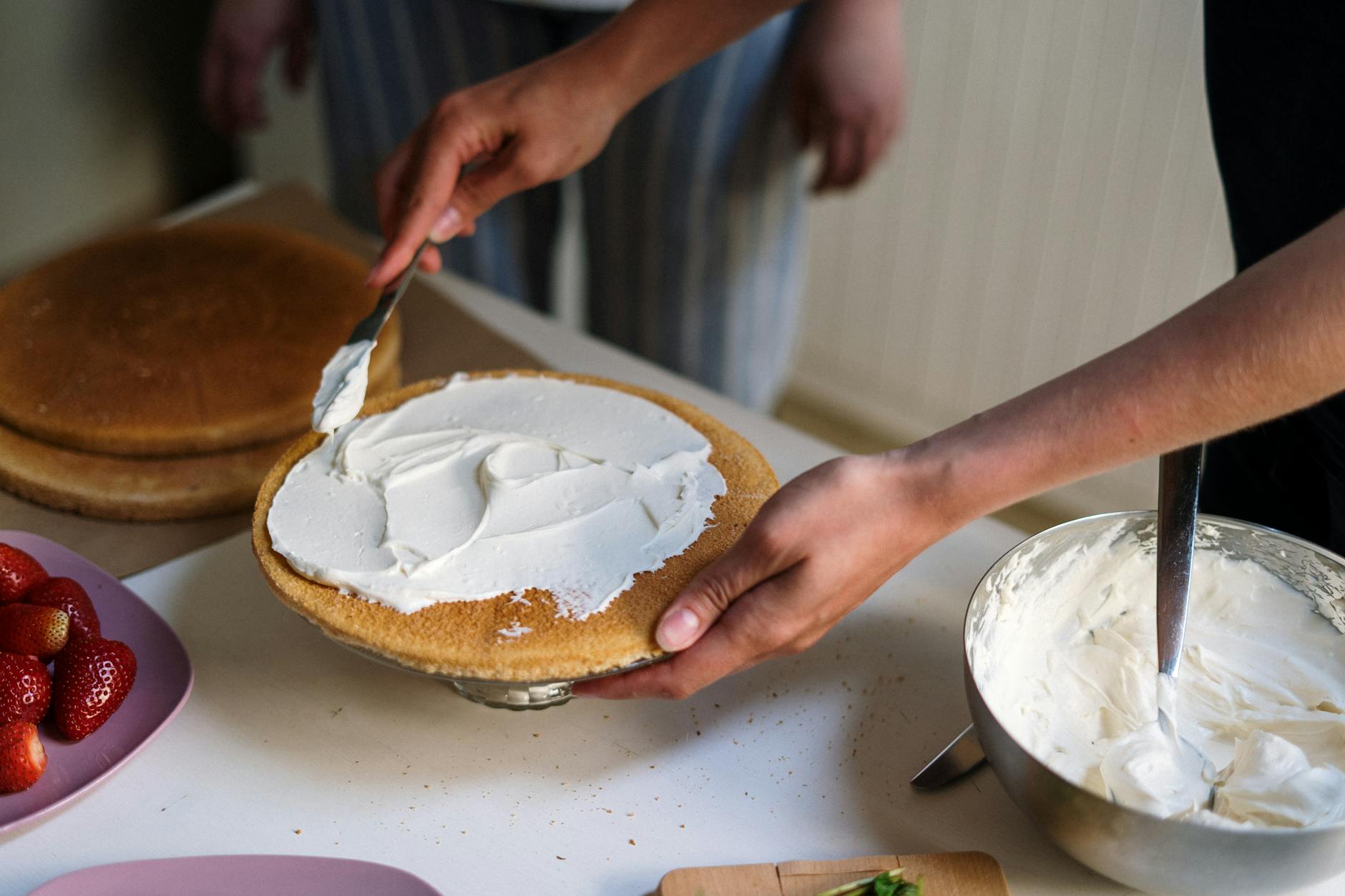Hands preparing a cake with cream frosting in a cozy kitchen setting, featuring strawberries and a mixing bowl.